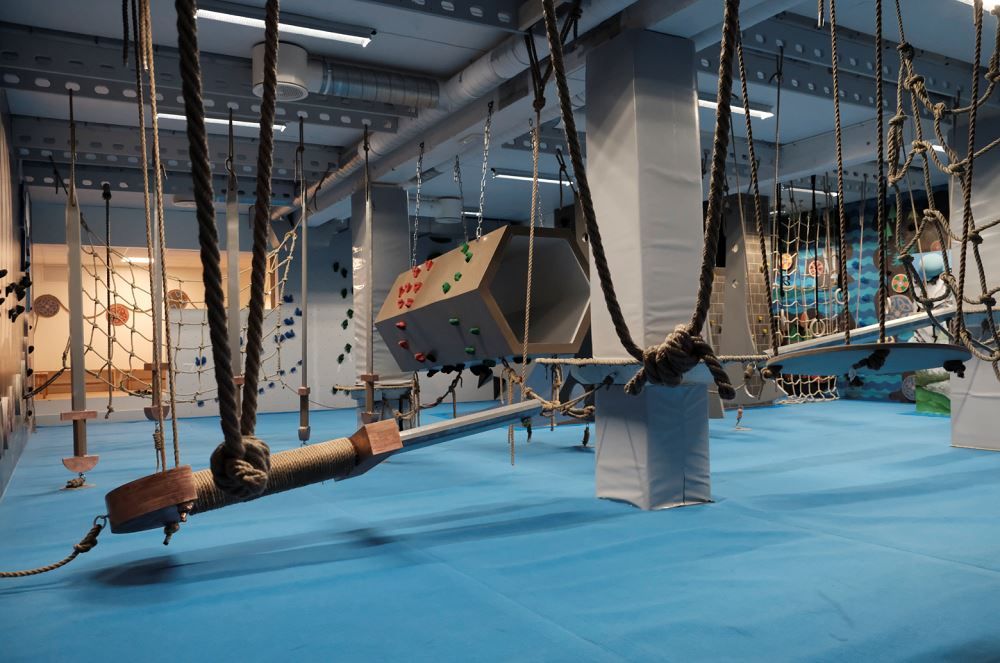 Indoor adventure gym with blue padded floor, hanging ropes and nets, and climbing obstacles under ceiling lights..