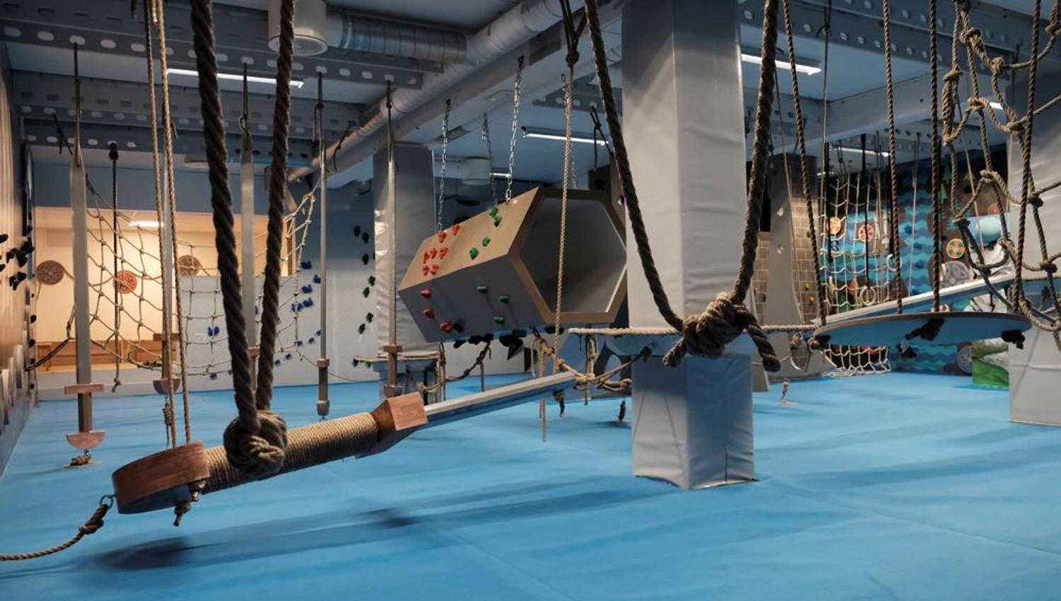 Indoor adventure gym with blue padded floor, hanging ropes and nets, and climbing obstacles under ceiling lights..