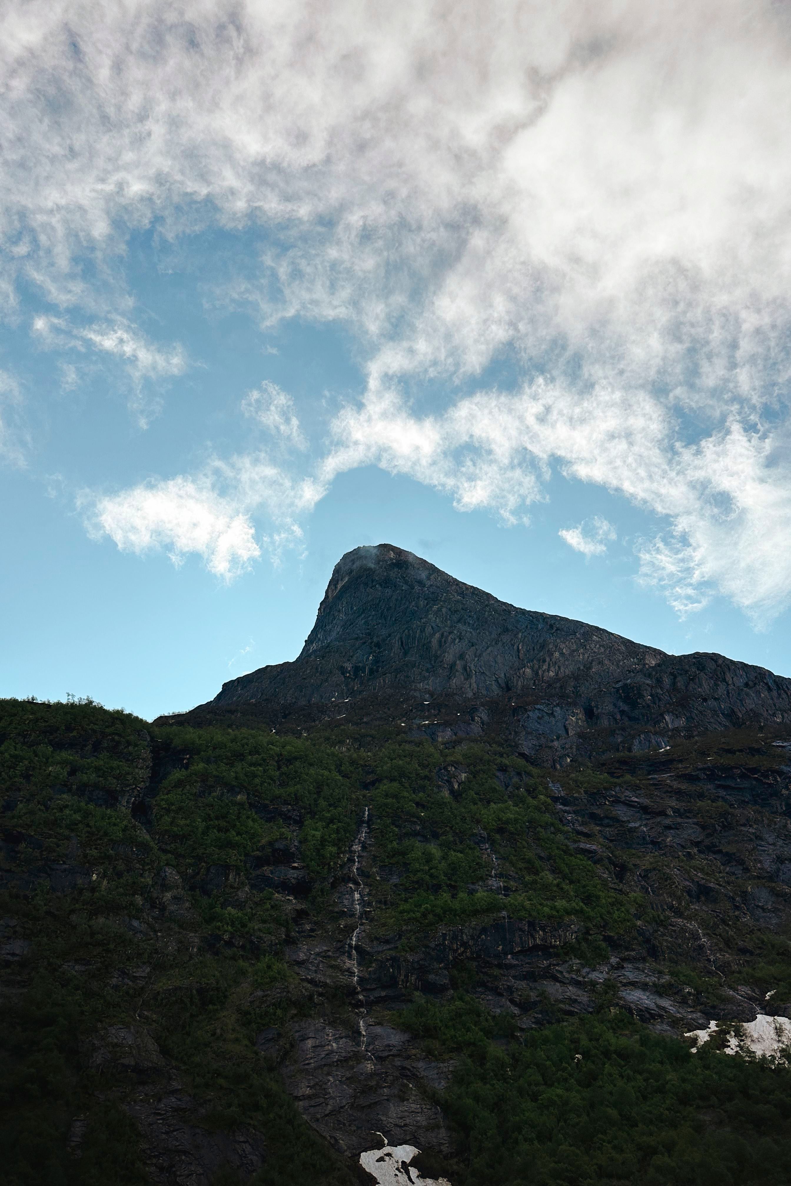 Low-angle view of a rocky mountain peak with green slopes beneath a blue sky filled with white clouds..