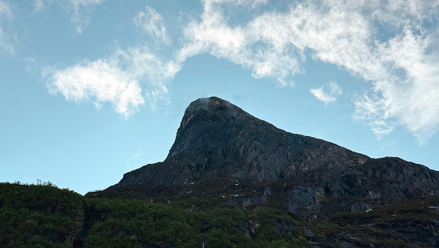 Low-angle view of a rocky mountain peak with green slopes beneath a blue sky filled with white clouds..