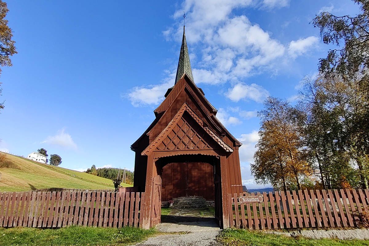 Tregjerde, portal med treskjæringer og stavkirke bakenfor. Blå himmel.