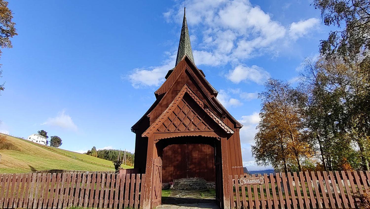 Tregjerde, portal med treskjæringer og stavkirke bakenfor. Blå himmel.