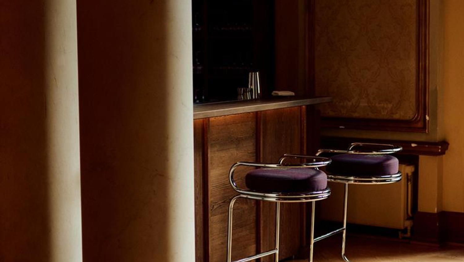 An elegant bar with dark wood, tall stools, and classical columns bathed in warm light.