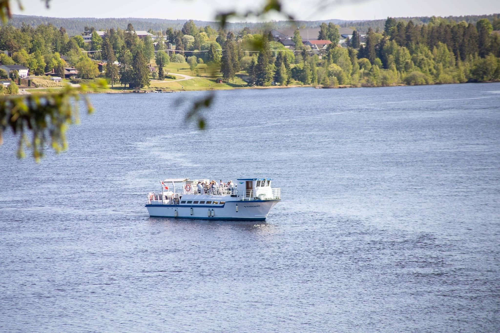 En liten, hvit rutebåt med mennesker om bord, som seiler på en elv omgitt av grønne trær i bakgrunnen.