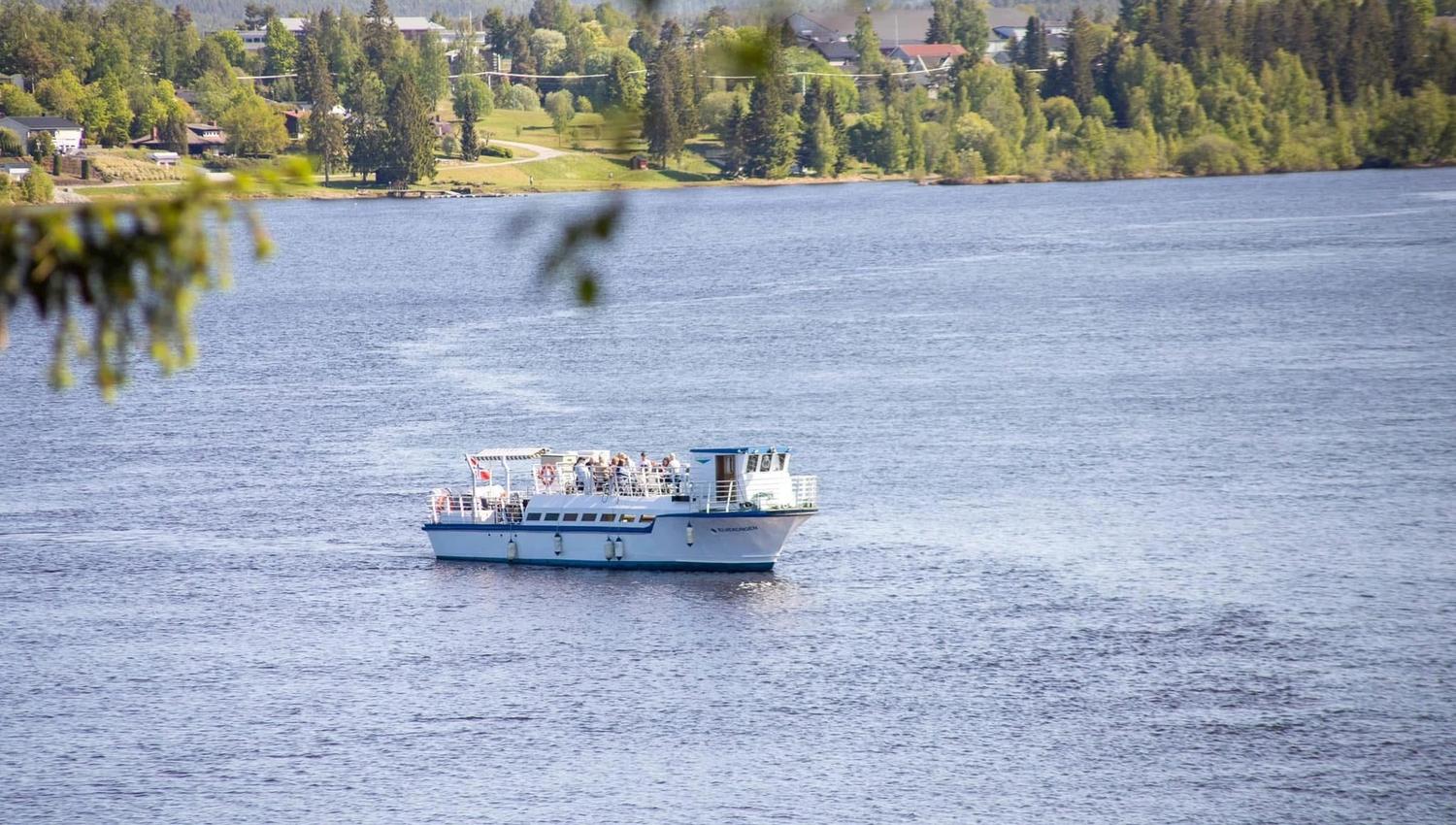 En liten, hvit rutebåt med mennesker om bord, som seiler på en elv omgitt av grønne trær i bakgrunnen.