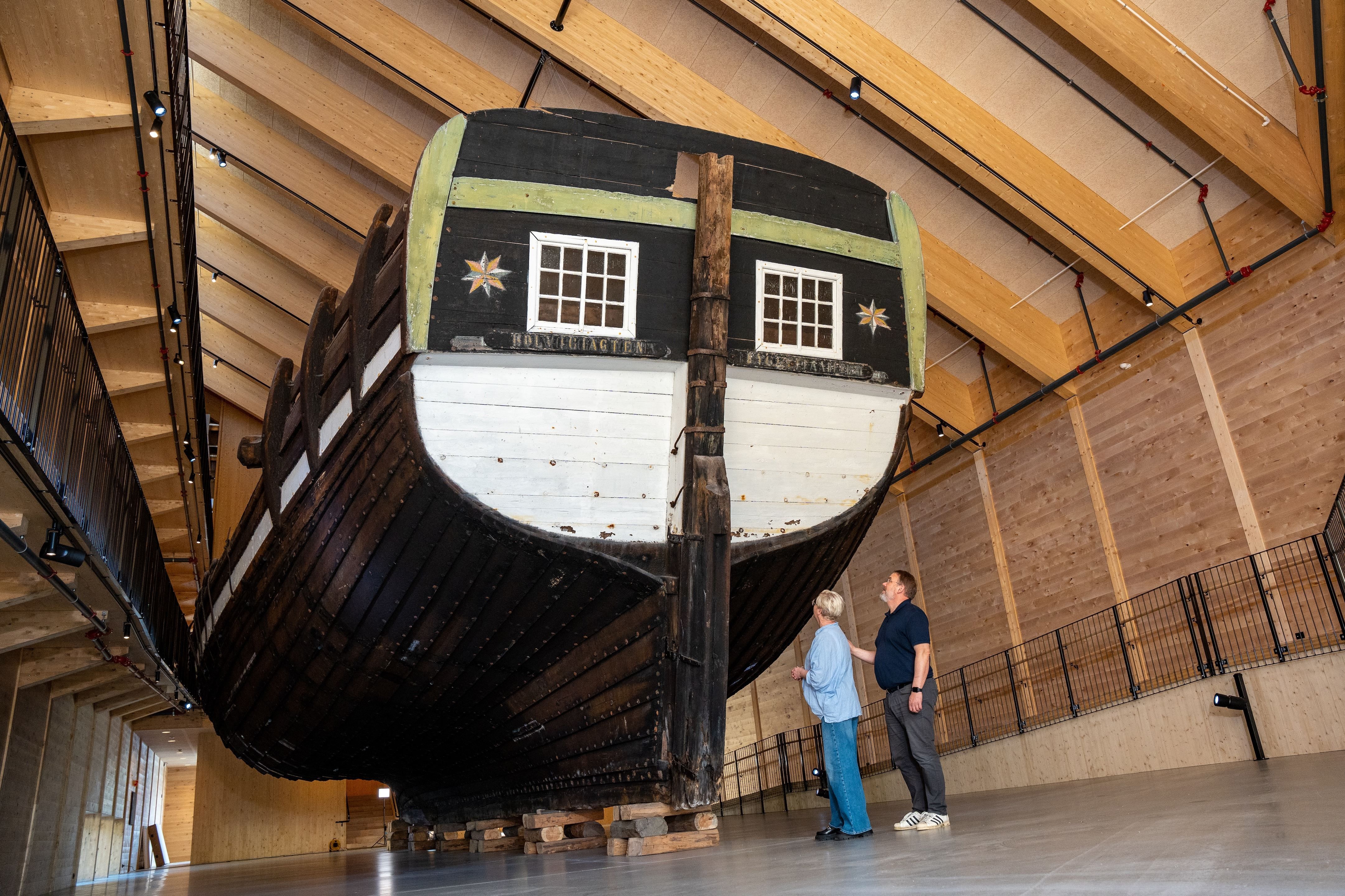 Large wooden ship, Holvikejekta, hull displayed inside a timber hall, with two visitors standing nearby on a concrete floor..