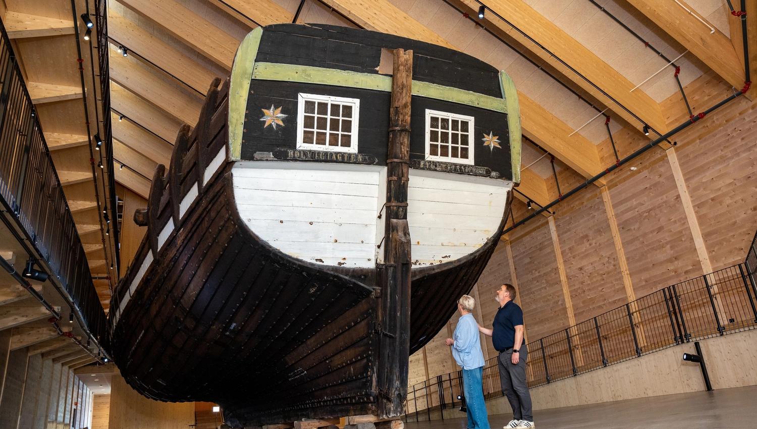 Large wooden ship, Holvikejekta, hull displayed inside a timber hall, with two visitors standing nearby on a concrete floor..