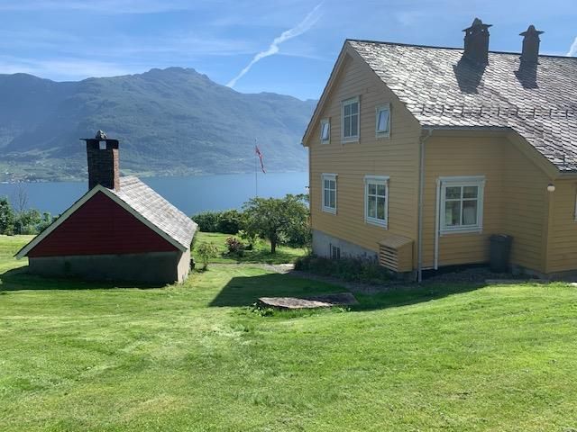 View from Jaastad Gjestetun featuring a yellow Swiss-style house, lush lawn, and panoramic views of the Hardangerfjord.