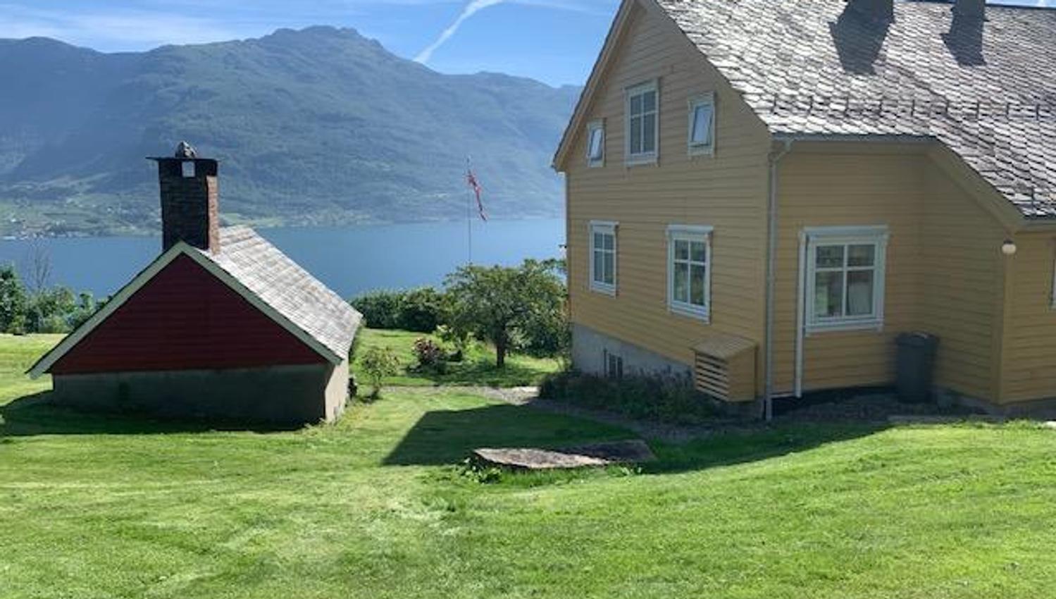 View from Jaastad Gjestetun featuring a yellow Swiss-style house, lush lawn, and panoramic views of the Hardangerfjord.