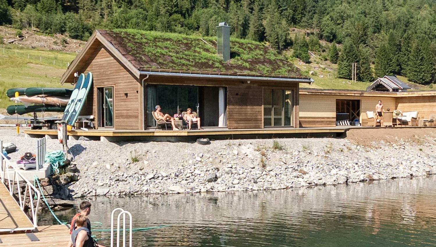 Wooden lakeside cabin with a green roof, people relaxing on a deck, kayaks nearby, and forested hills behind..