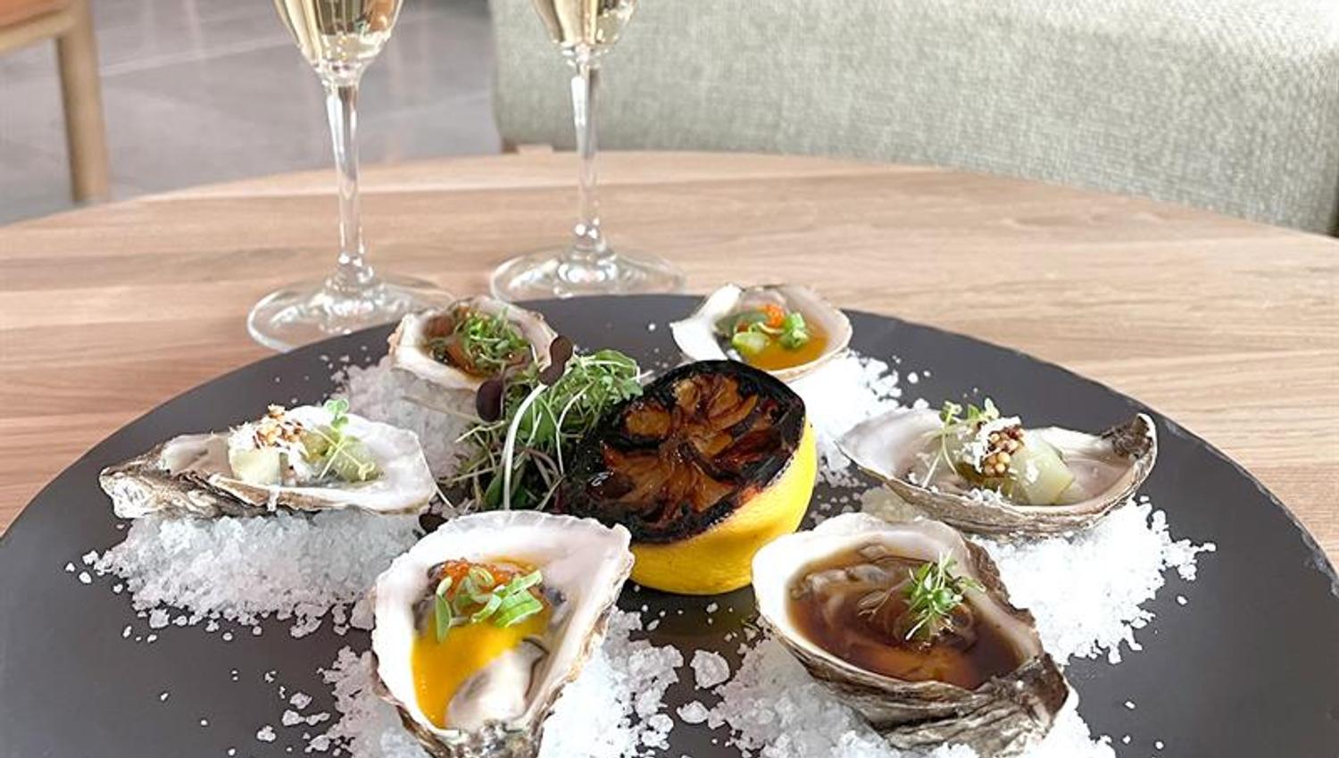 Detail of oyster on a plate with salt sprinkled around and half a lemon in the middle. Two glasses of sparkling white wine next to it.