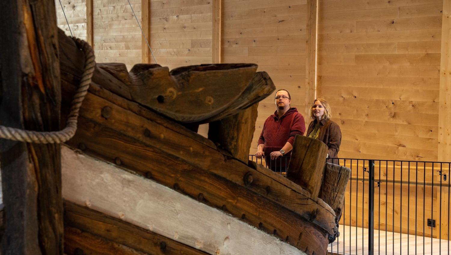 Large wooden ship bow on display indoors, with two visitors standing behind a railing against wood-paneled walls..