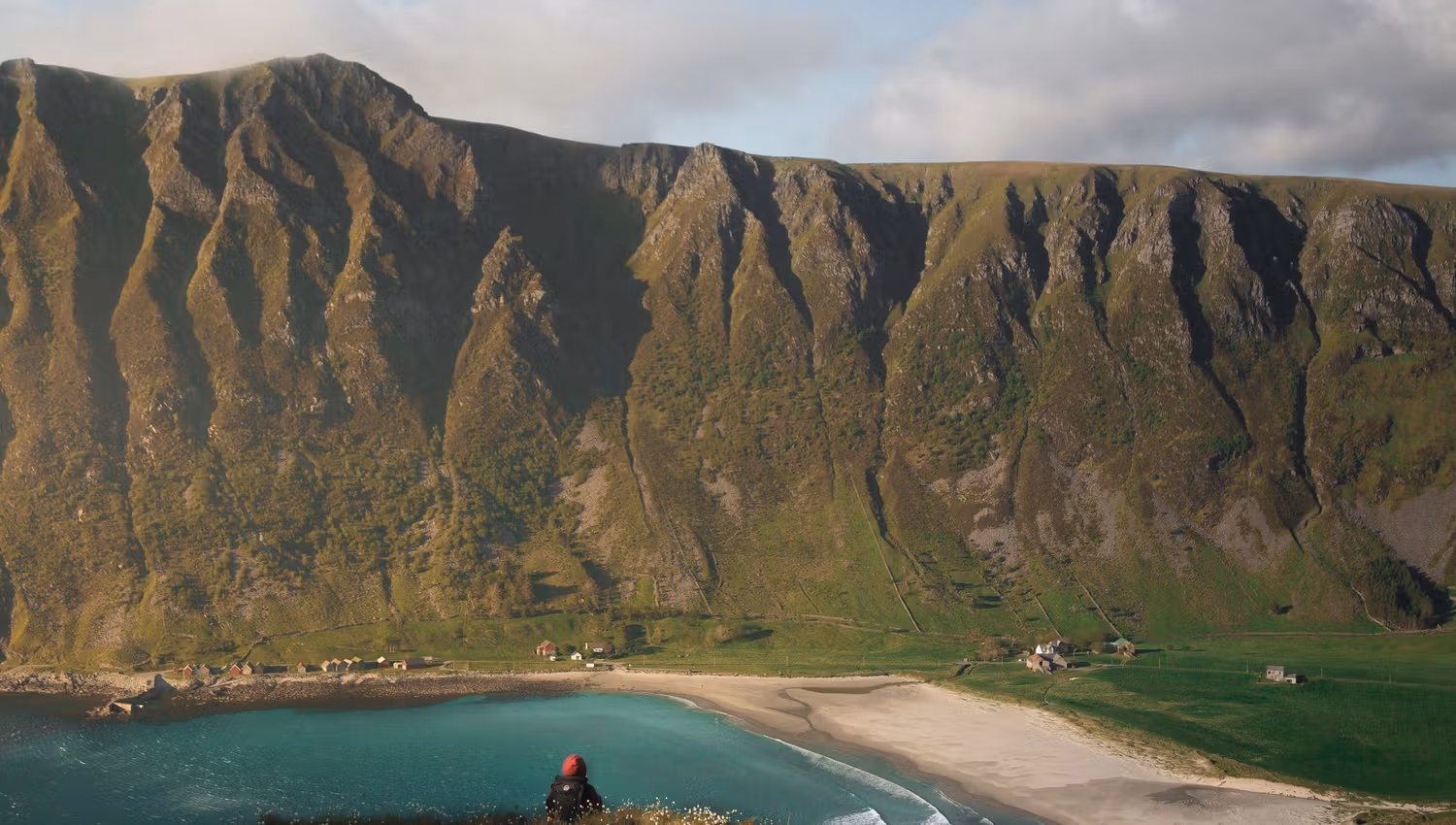 Ein person sit på ein kolle og ser ned mot turkis hav, sandstrand, grøne marker og bratte fjell under skyer..