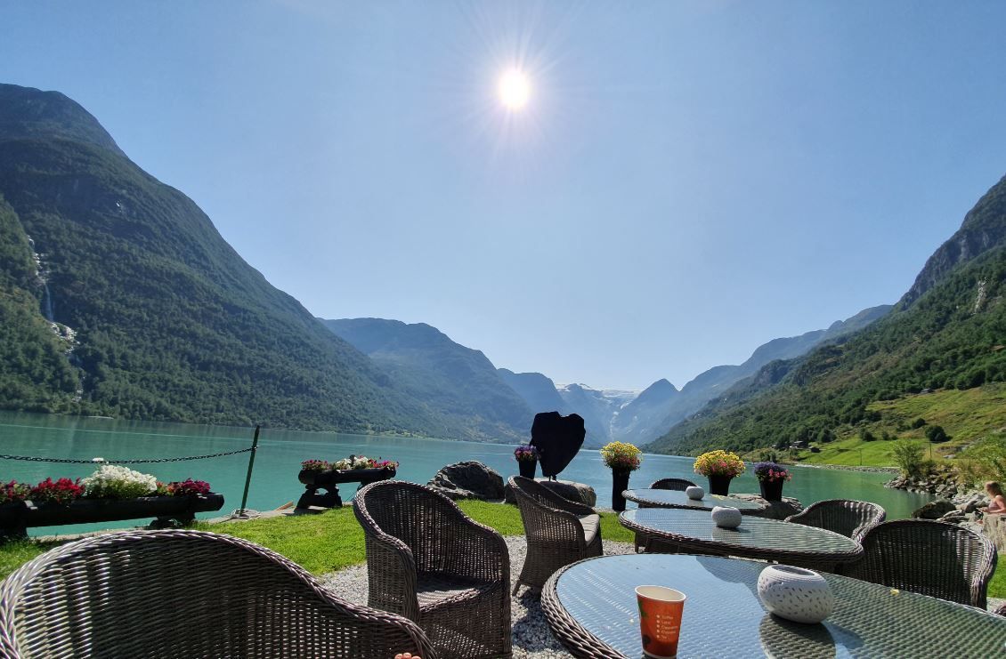Outdoor patio with wicker chairs and glass tables beside a turquoise lake, surrounded by green mountains under sun.