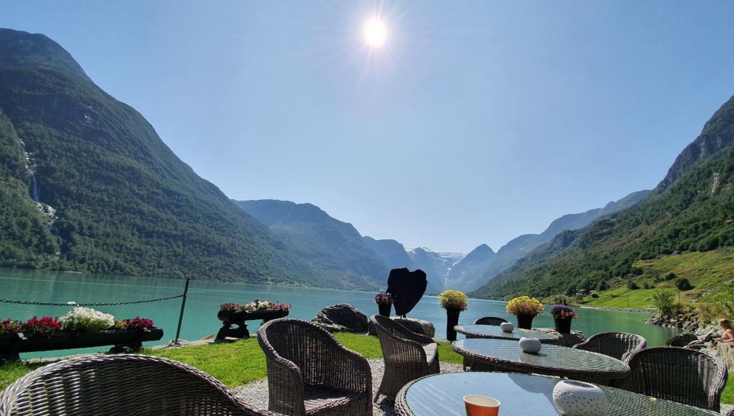 Outdoor patio with wicker chairs and glass tables beside a turquoise lake, surrounded by green mountains under sun.