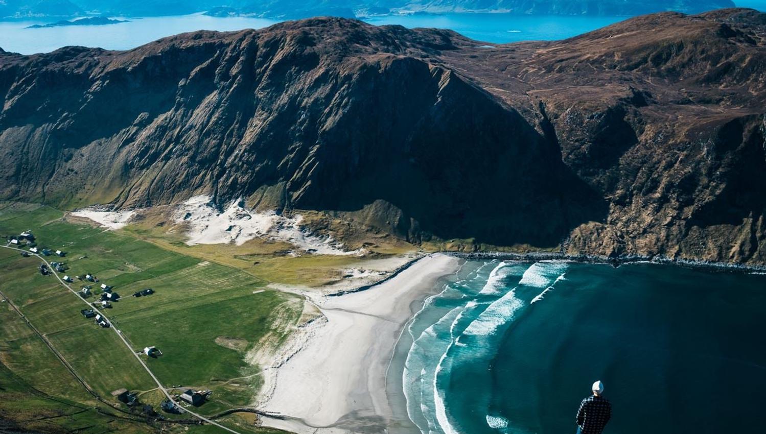 Ein person står på ein fjelltopp og ser ned på ei kvit sandstrand, bølgjer, grøne jorder og bratte fjell ved fjorden..