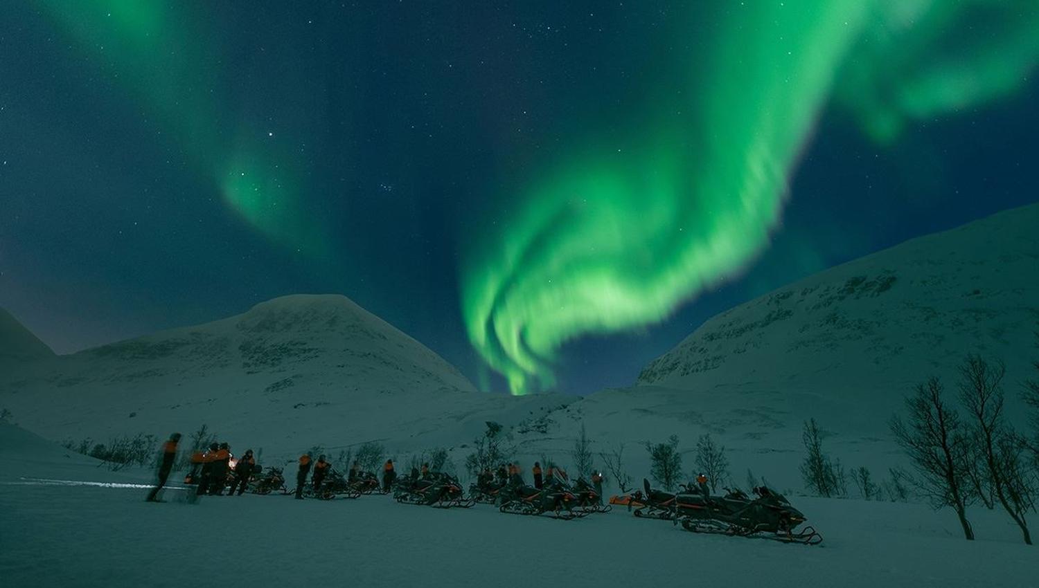 Gjester står ved snøskuterne og beundrer det vakre nordlyset