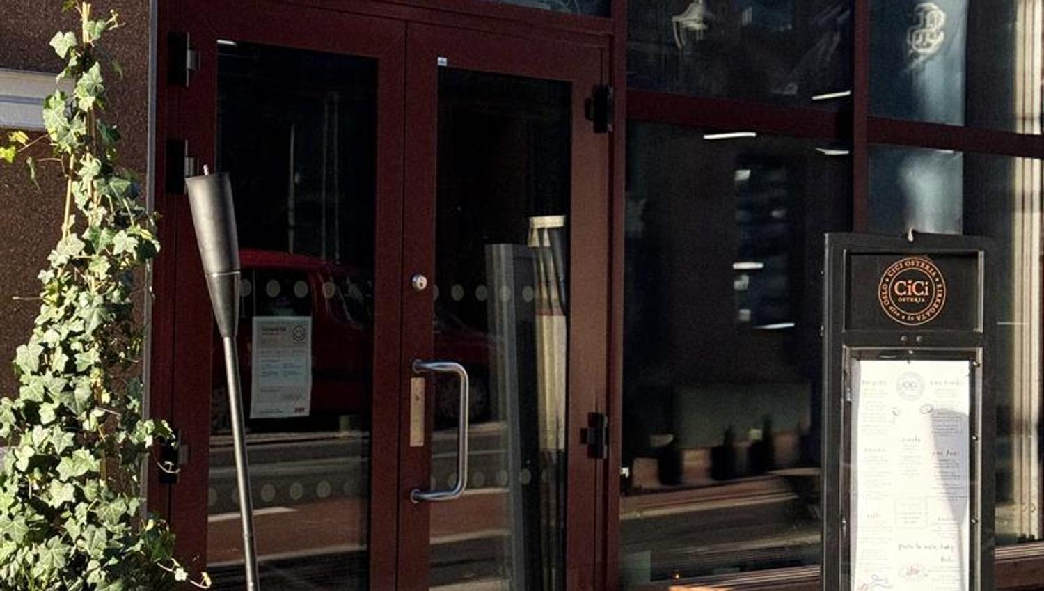 Inngangspartiet til en restaurant med store glassvinduer, menyplakat ved døren og en lykt og plante plassert på fortauet.