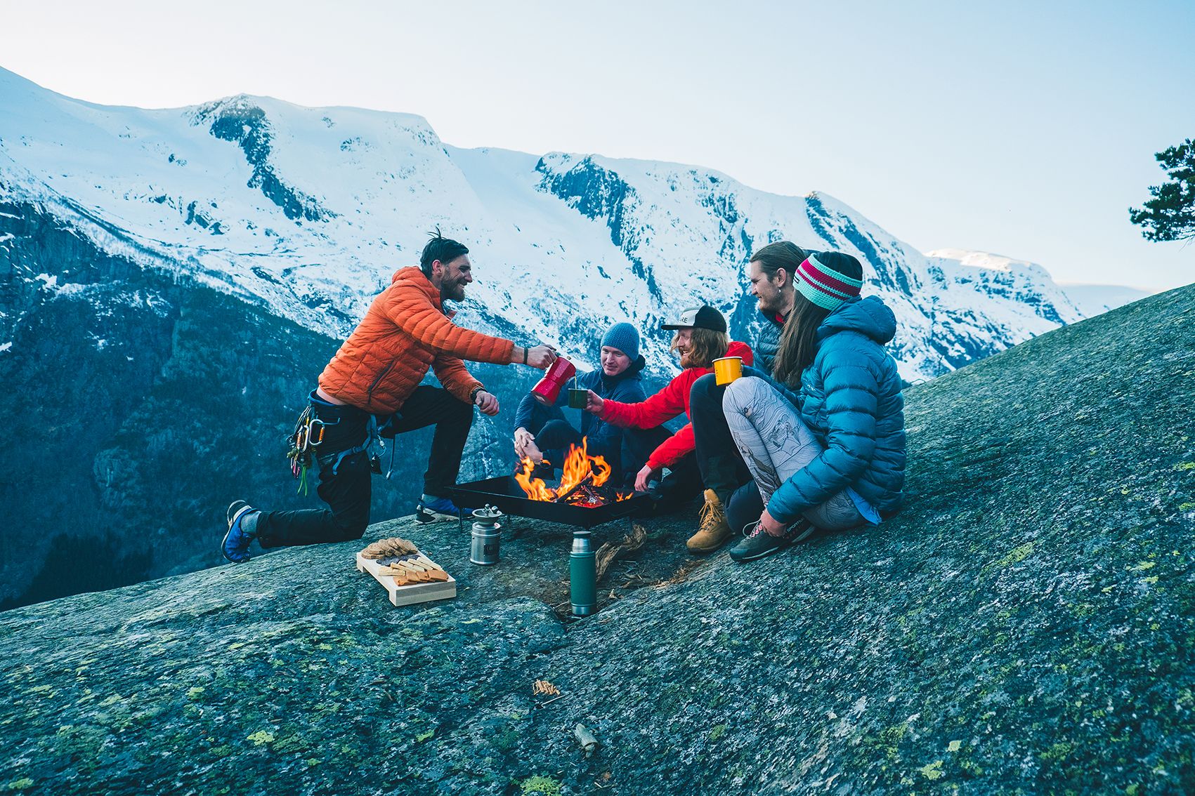 Ei gruppe venner kosar seg rundt eit bål på fjellet med utsikt over snødekte toppar i Hardanger. Sosialt samvær i spektakulære omgjevnader.