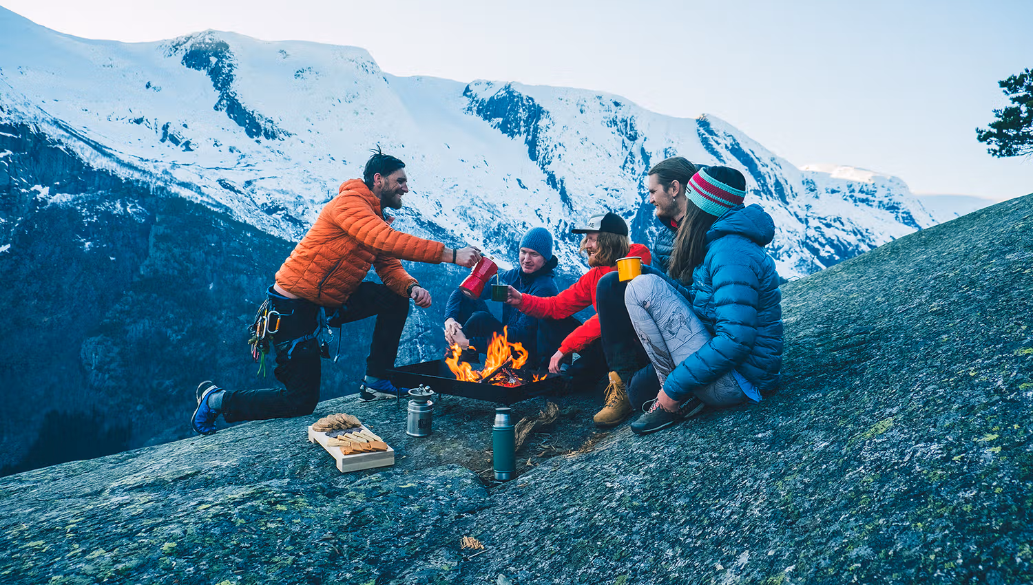 Ei gruppe venner kosar seg rundt eit bål på fjellet med utsikt over snødekte toppar i Hardanger. Sosialt samvær i spektakulære omgjevnader.