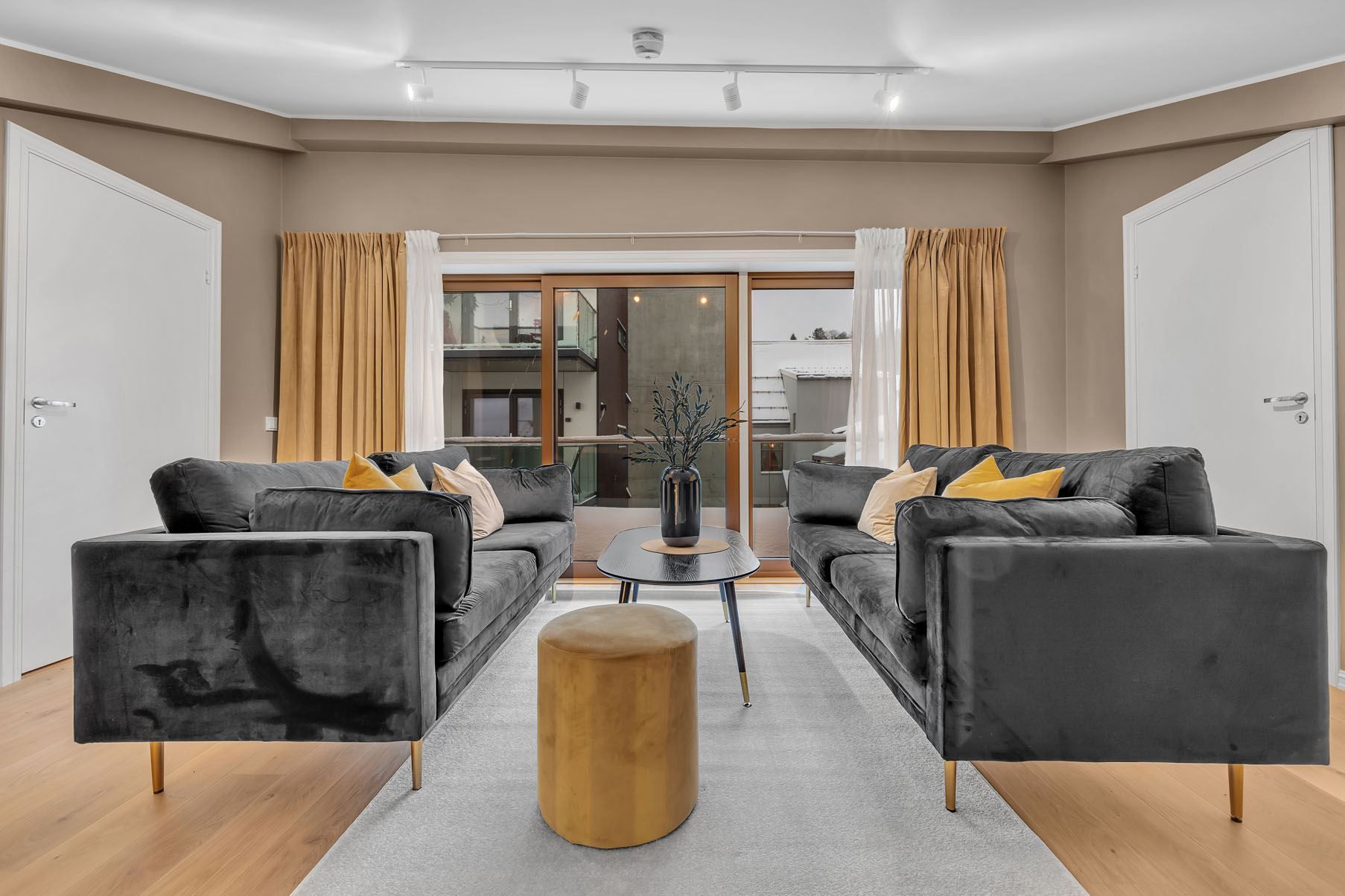 Stue med mørkegrå sofaer, lyst tregulv og store glassdører med gardiner.