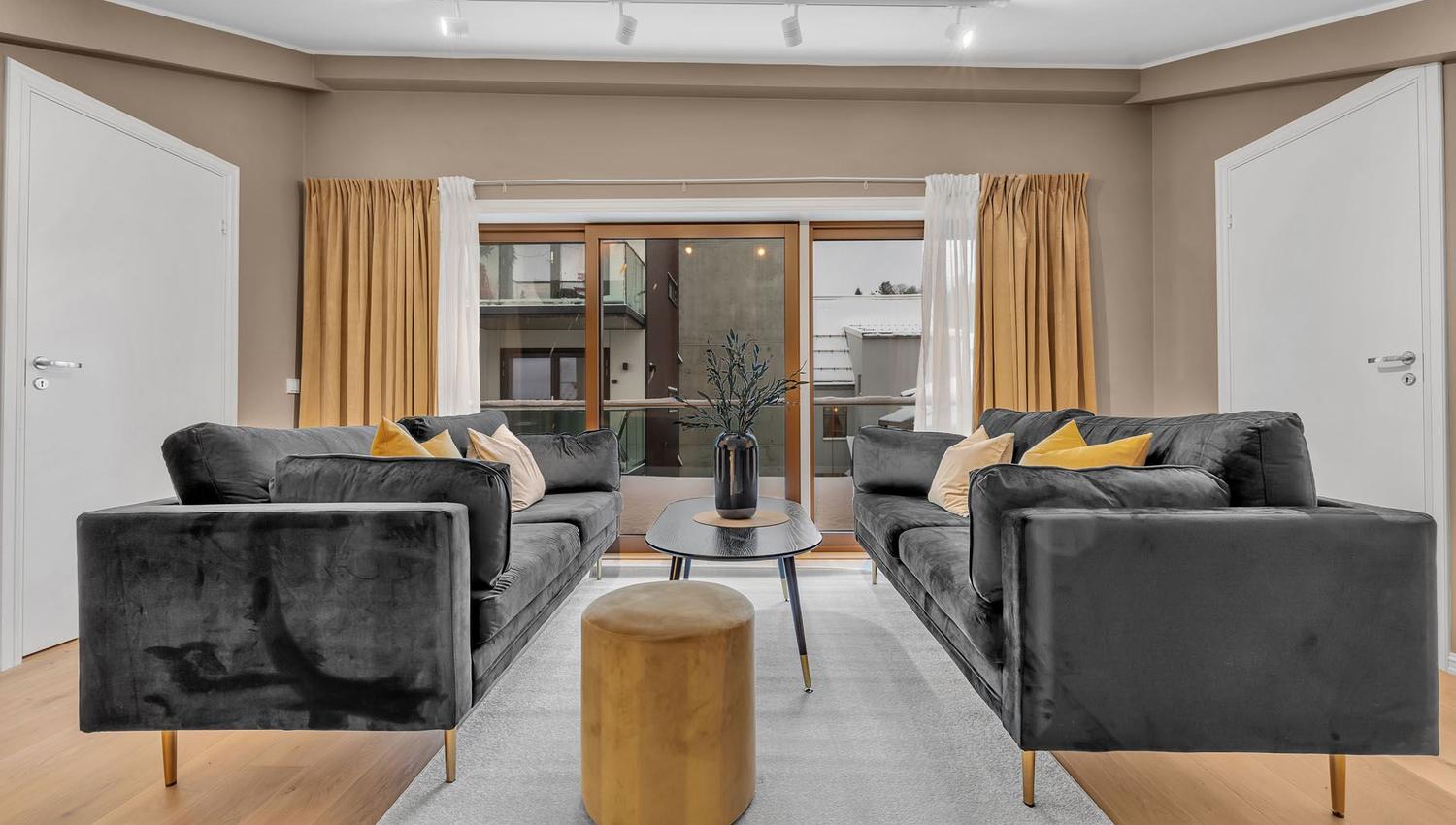 Stue med mørkegrå sofaer, lyst tregulv og store glassdører med gardiner.