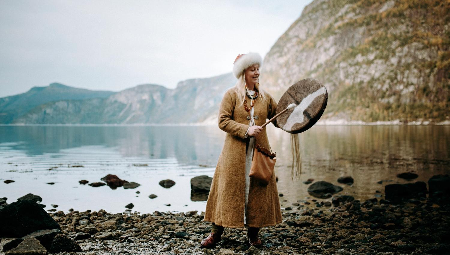 Eine als Wikingerin verkleidete Frau steht am Fjord und spielt eine Wikingertrommel.
