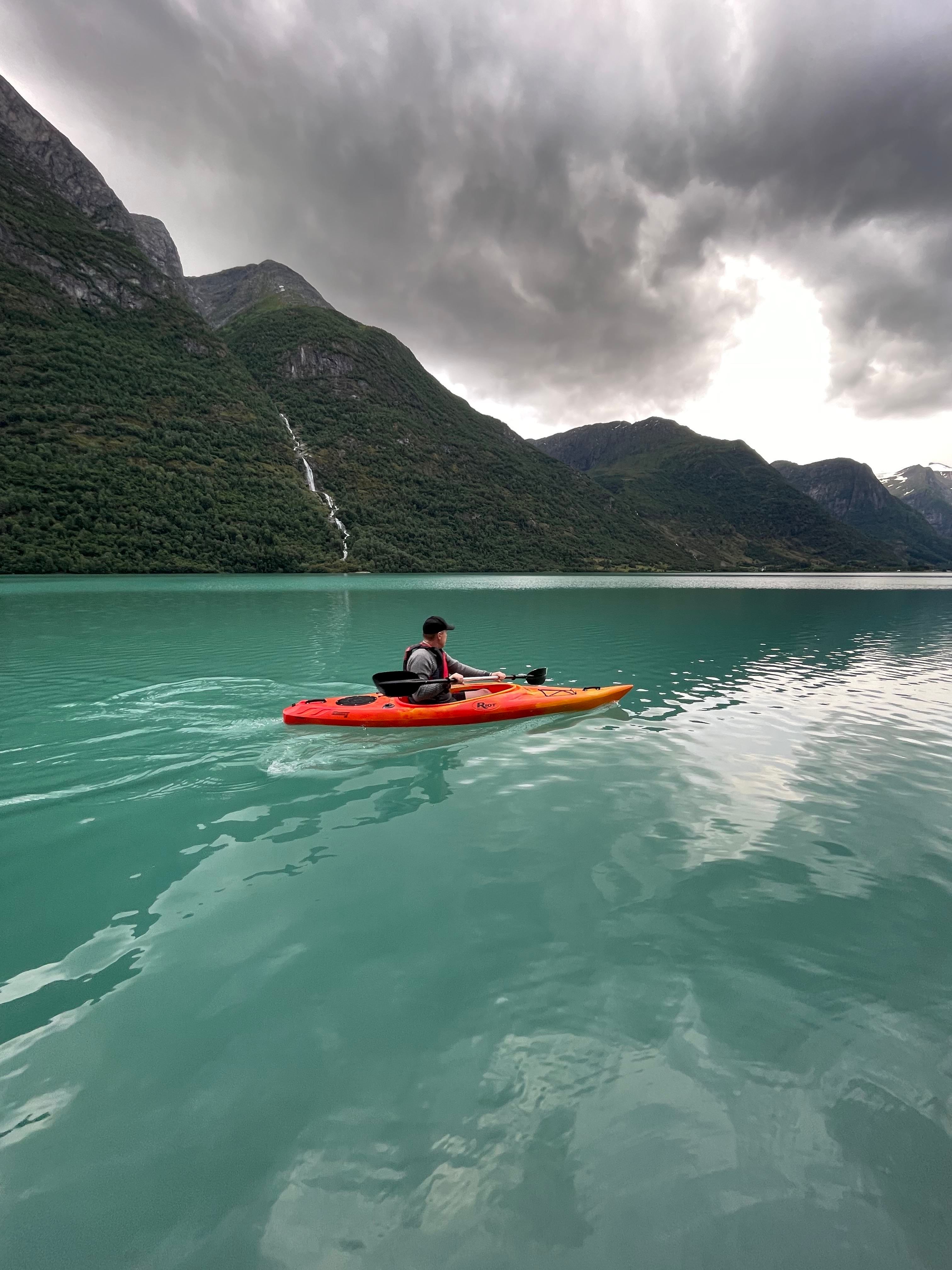 A person paddles an orange kayak on a turquoise lake, with green mountains and dark storm clouds overhead..