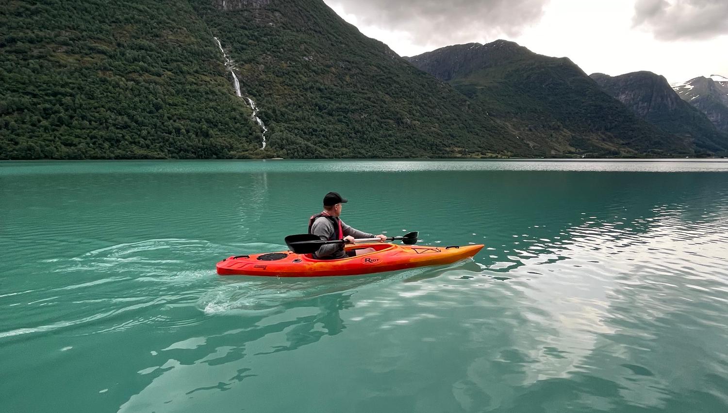 A person paddles an orange kayak on a turquoise lake, with green mountains and dark storm clouds overhead..