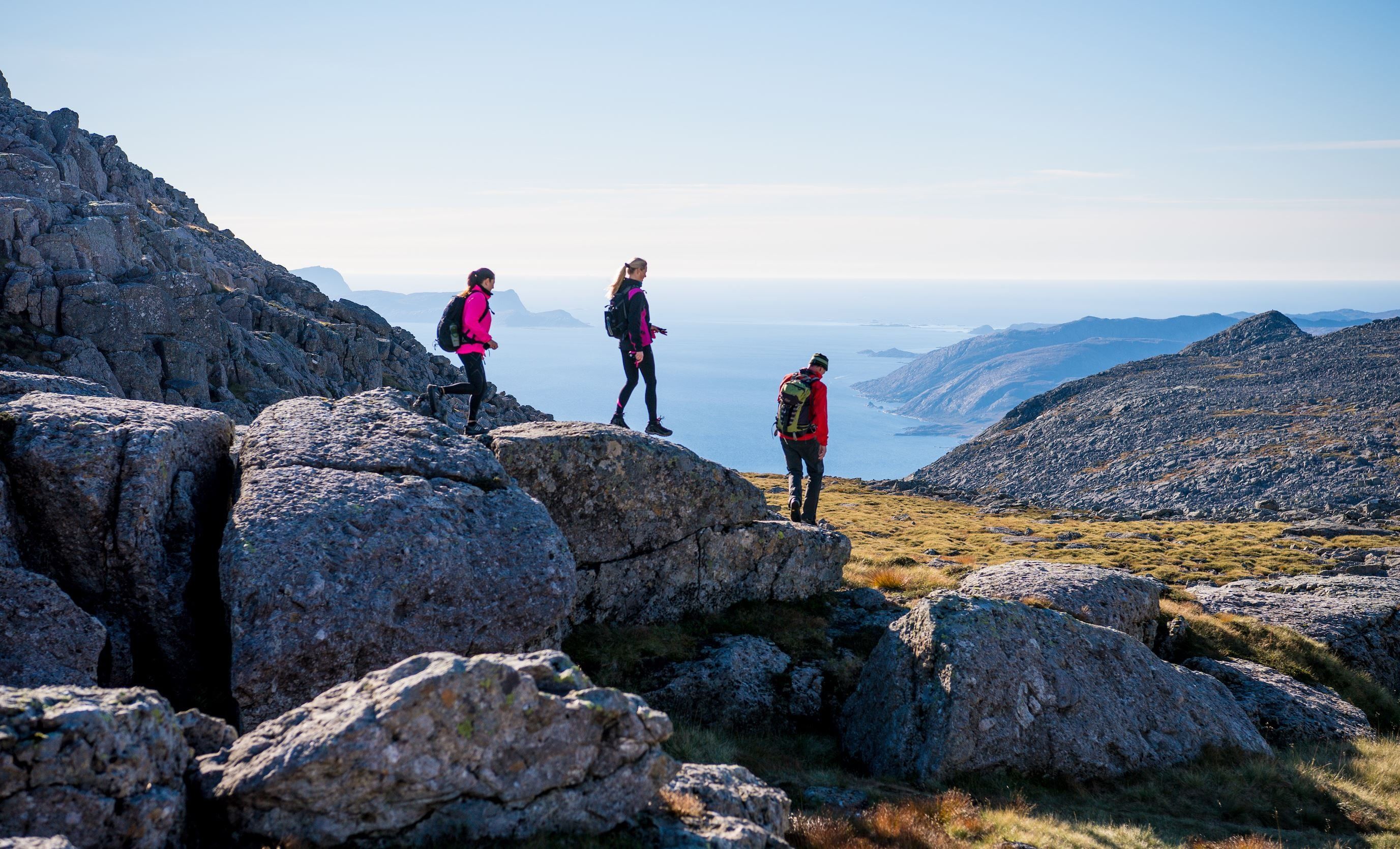 Tre turgåarar med ryggsekk står på steinur i fjellet og ser ut mot hav og fjord under klar himmel..