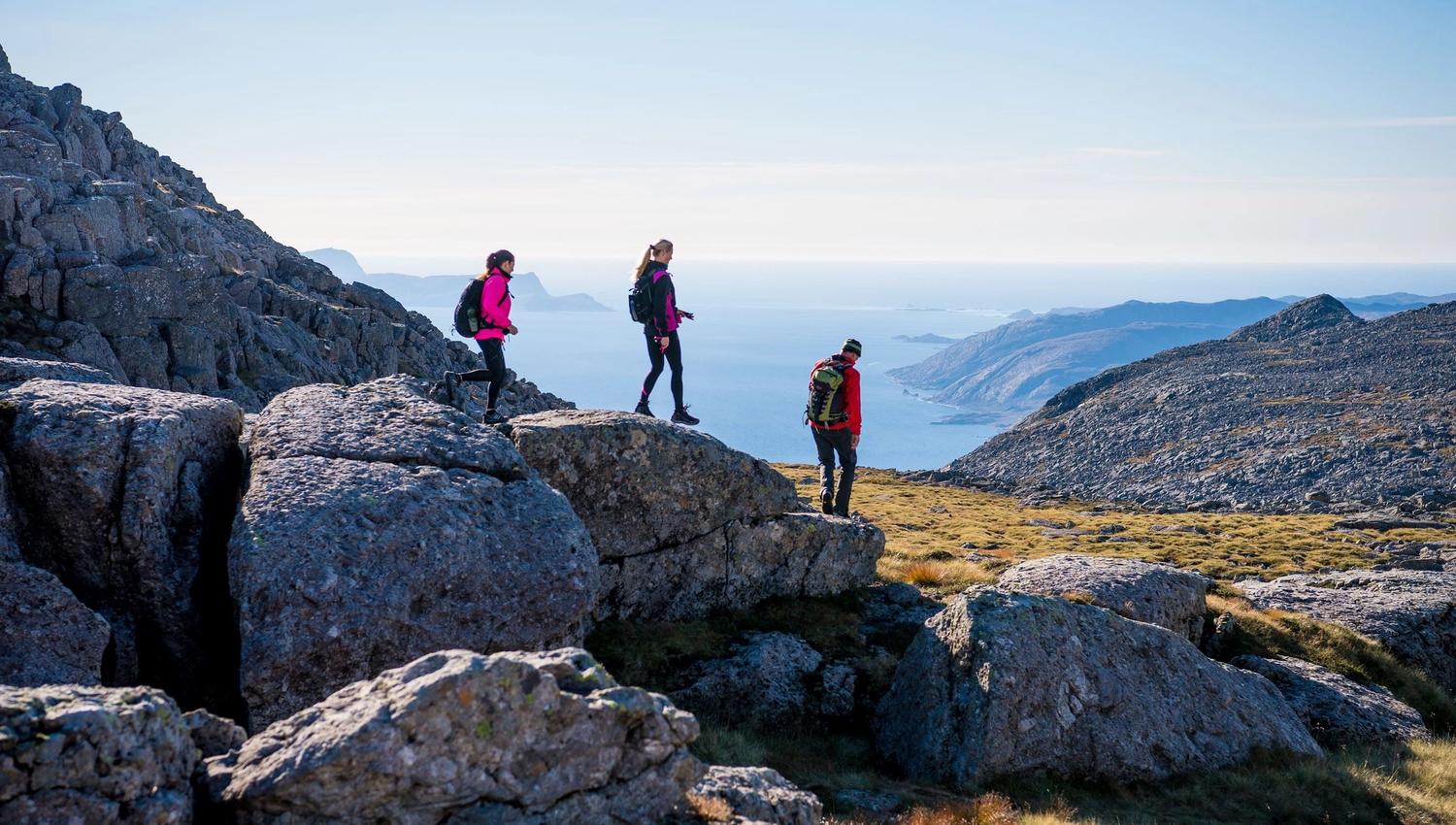 Tre turgåarar med ryggsekk står på steinur i fjellet og ser ut mot hav og fjord under klar himmel..