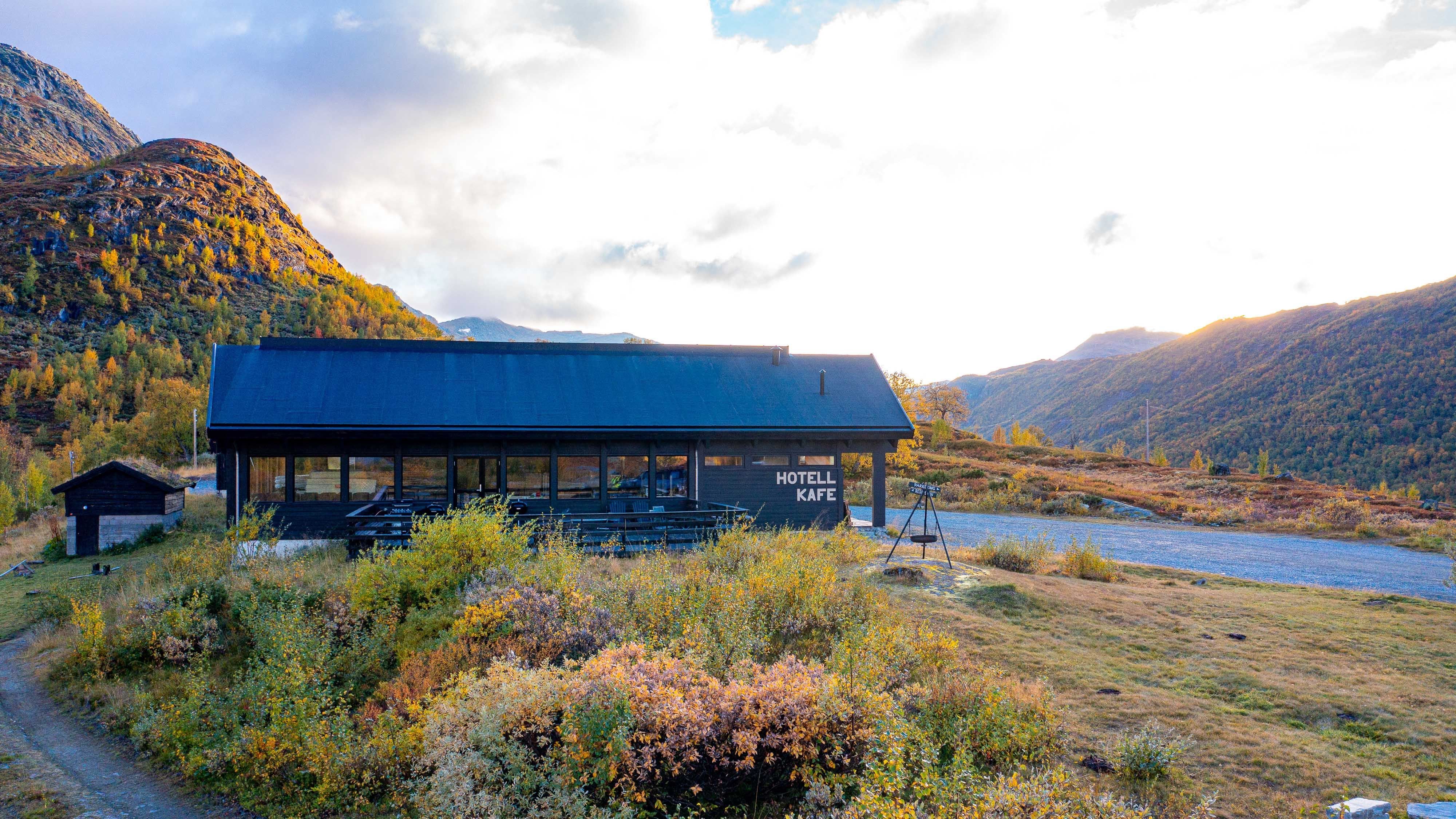 Een donkere houten lodge met grote ramen ligt in een bergvallei, omringd door herfststruiken en heuvels onder wolken..