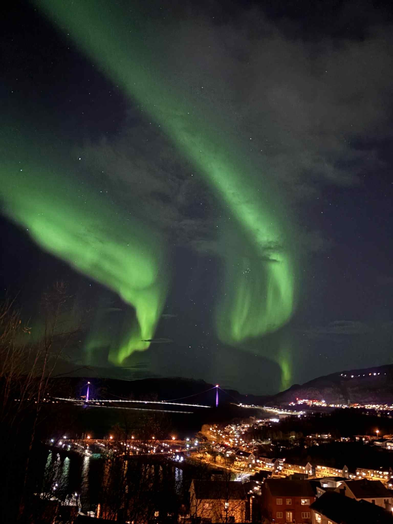 Green aurora borealis swirls across a cloudy night sky above a lit town, bridge, mountains, and water.