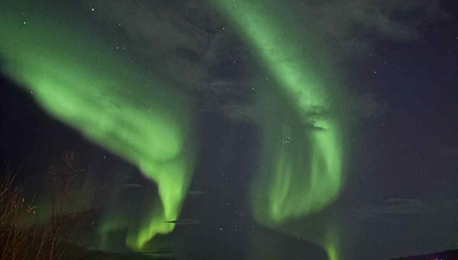 Green aurora borealis swirls across a cloudy night sky above a lit town, bridge, mountains, and water.
