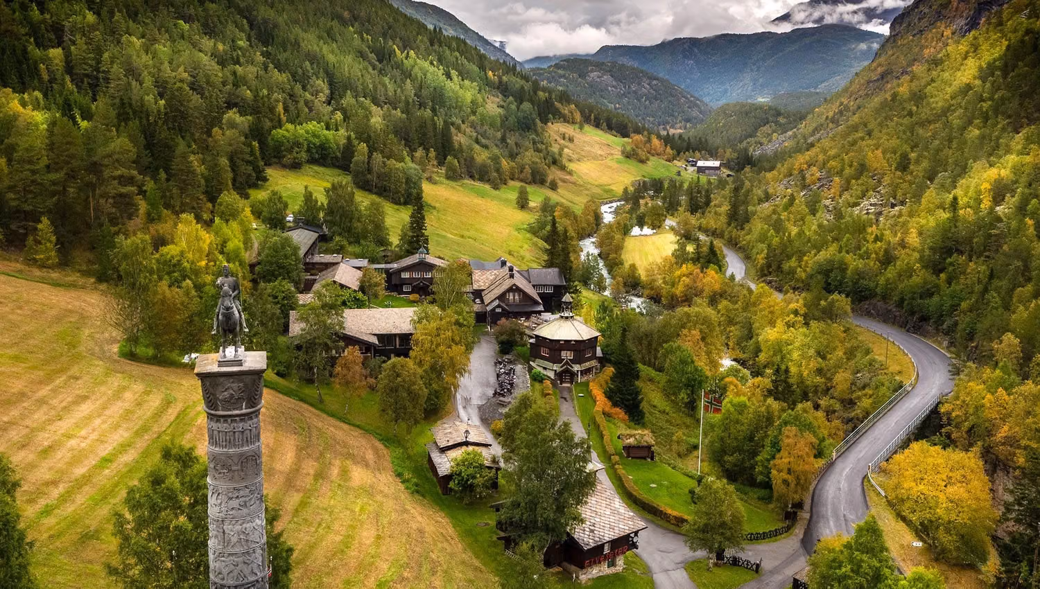 Landskap med grøne fjellsider, gardstun, svingete veg og elv i dalen, under skyer med fjell i bakgrunnen..