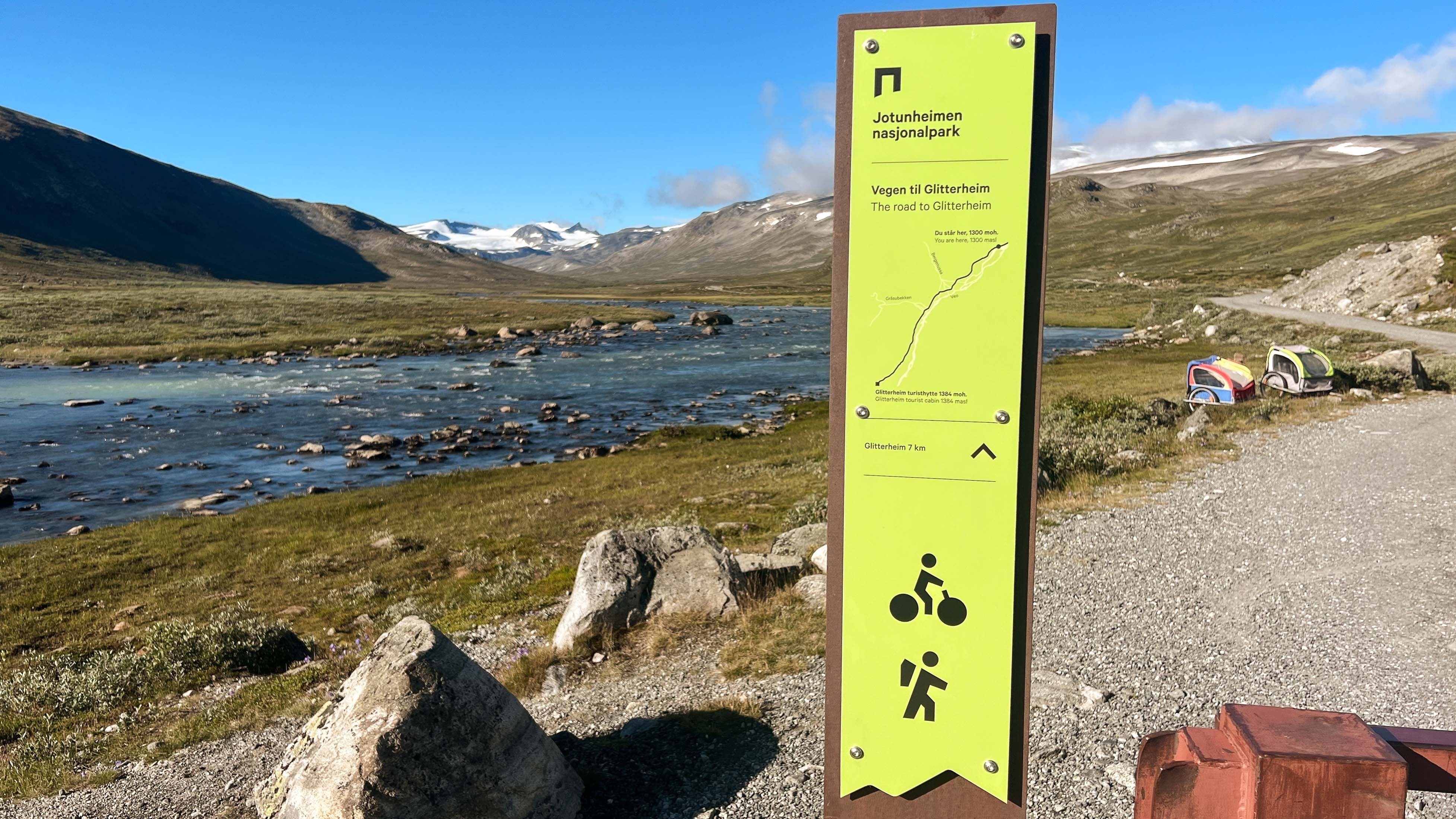 Green trail sign beside a gravel path and river in a wide mountain valley with distant snow-capped peaks..