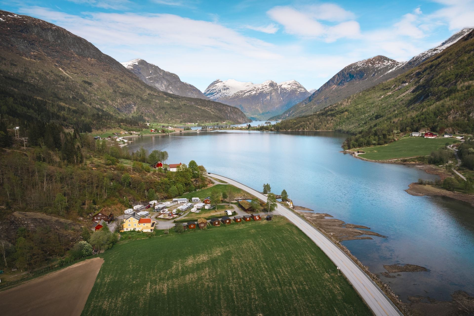 A wide valley lake bordered by green fields and small buildings, with forested slopes and snowy mountains beyond..