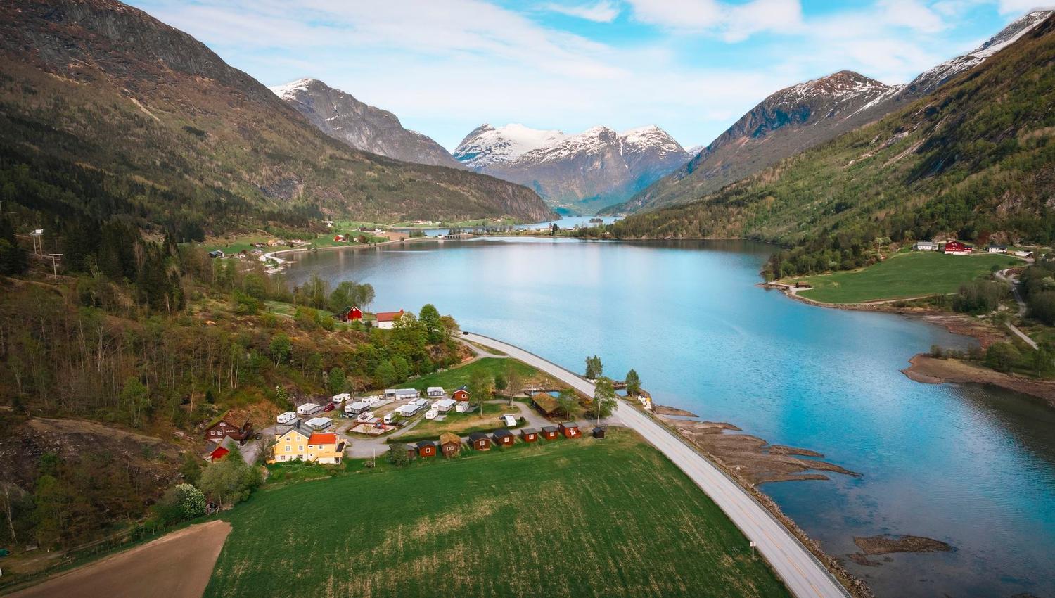 A wide valley lake bordered by green fields and small buildings, with forested slopes and snowy mountains beyond..