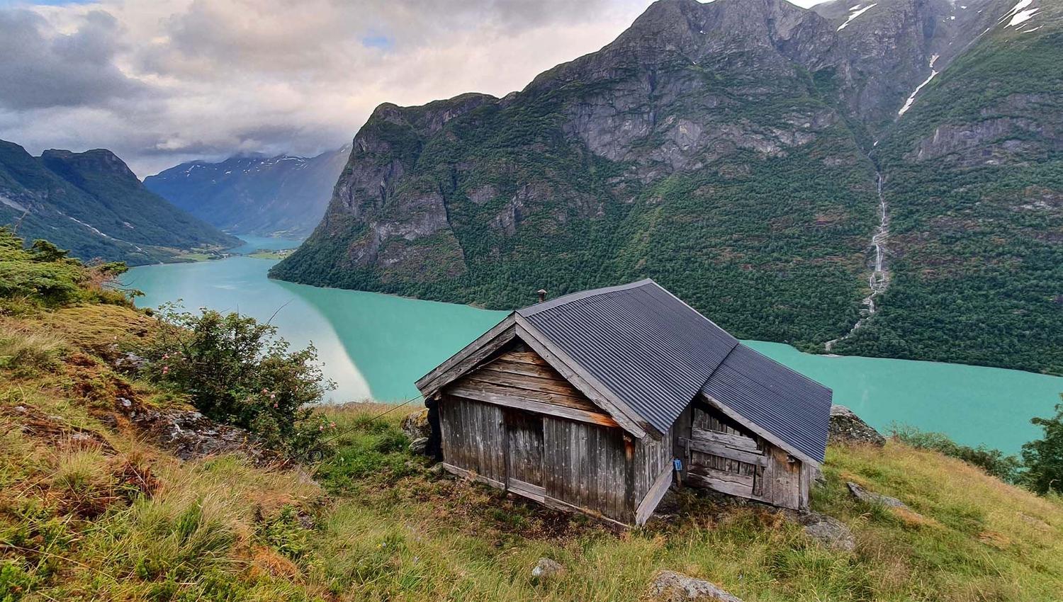 Weathered wooden cabin on a grassy hillside above a turquoise fjord, with steep forested mountains and clouds.