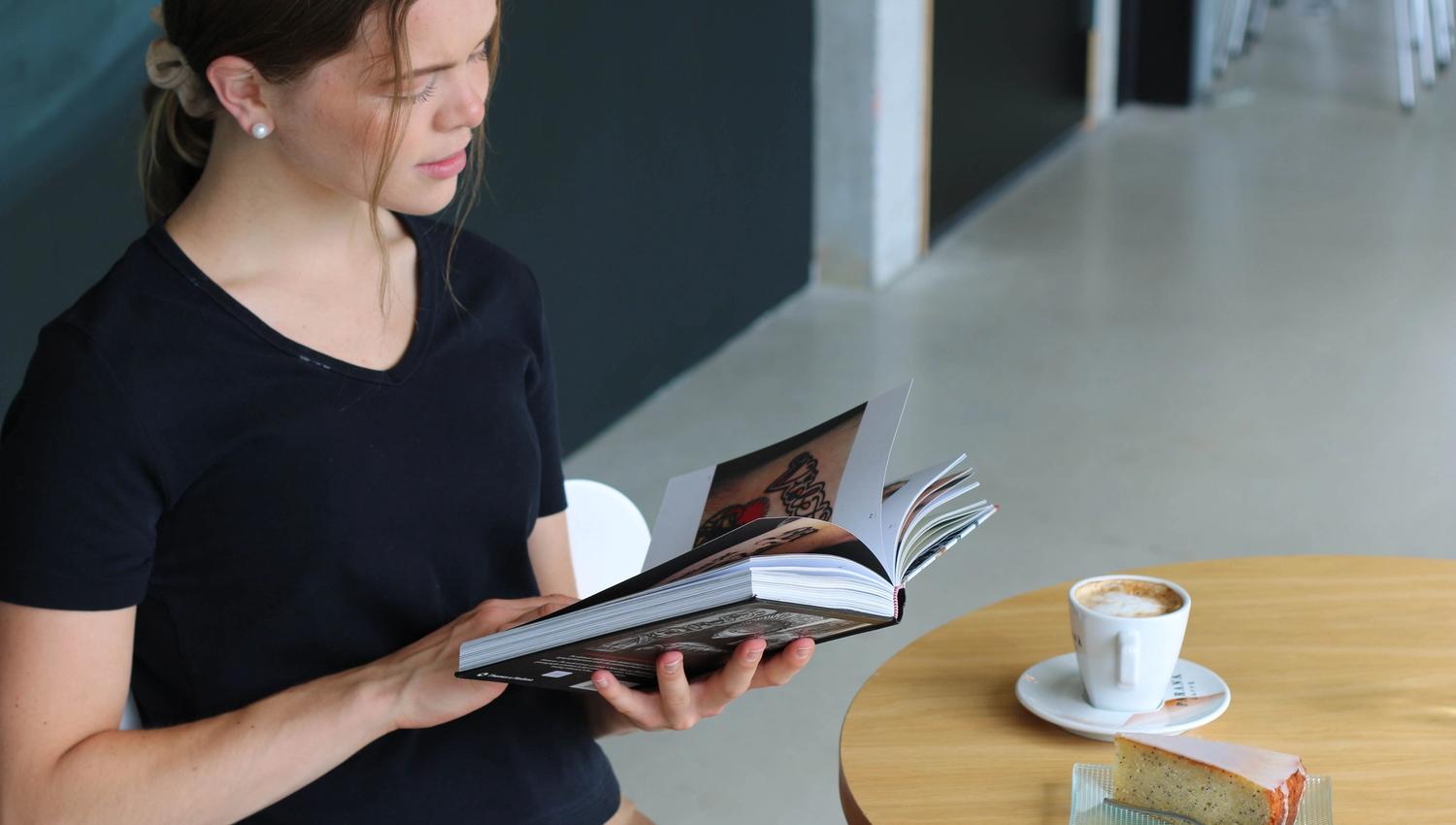 A young woman sits at a table reading a book, with a cup of coffee and a slice of cake placed in front of her.