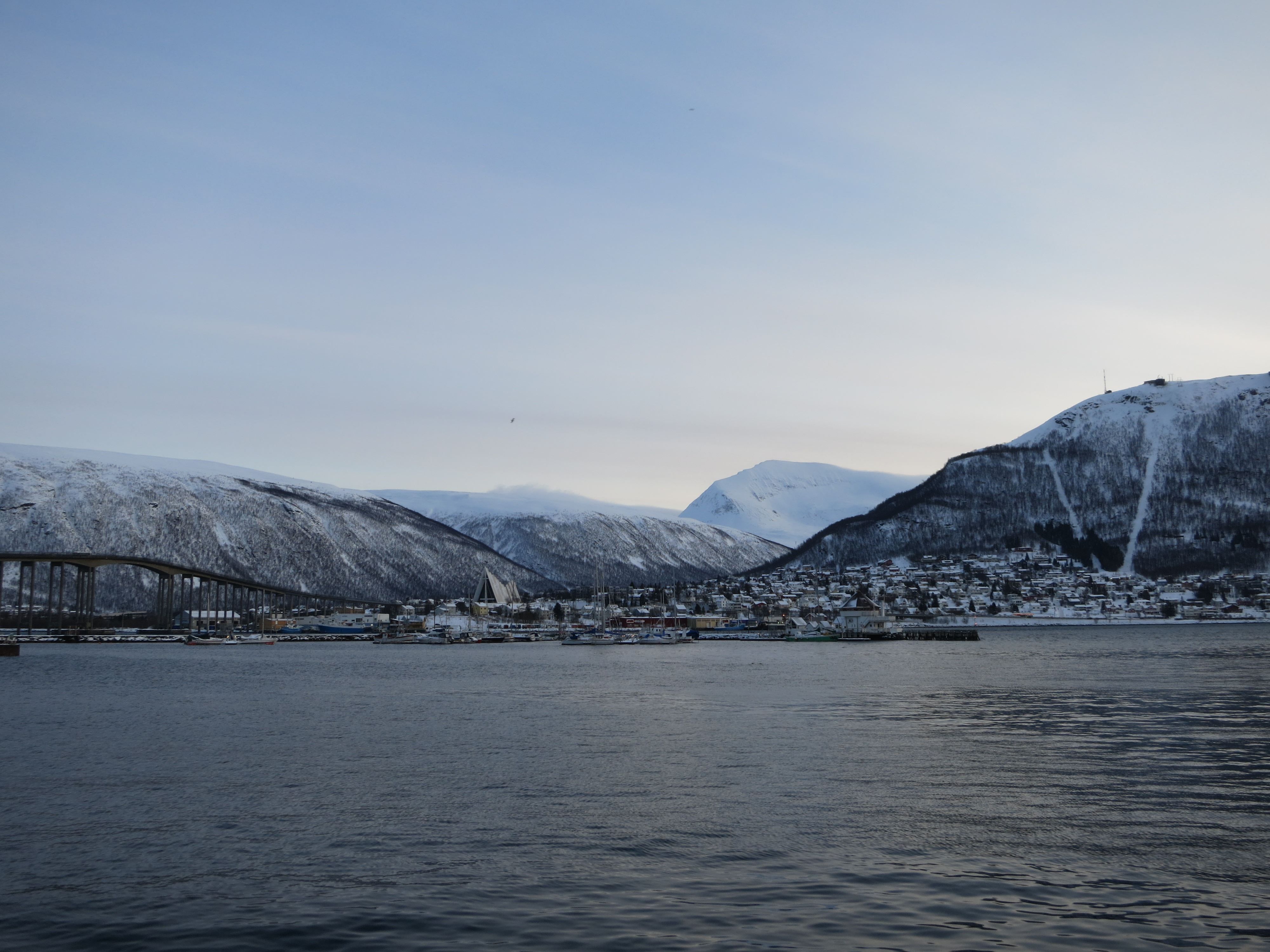 Tromsø with a bridge, buildings, and snow-covered mountains reflected in calm water.