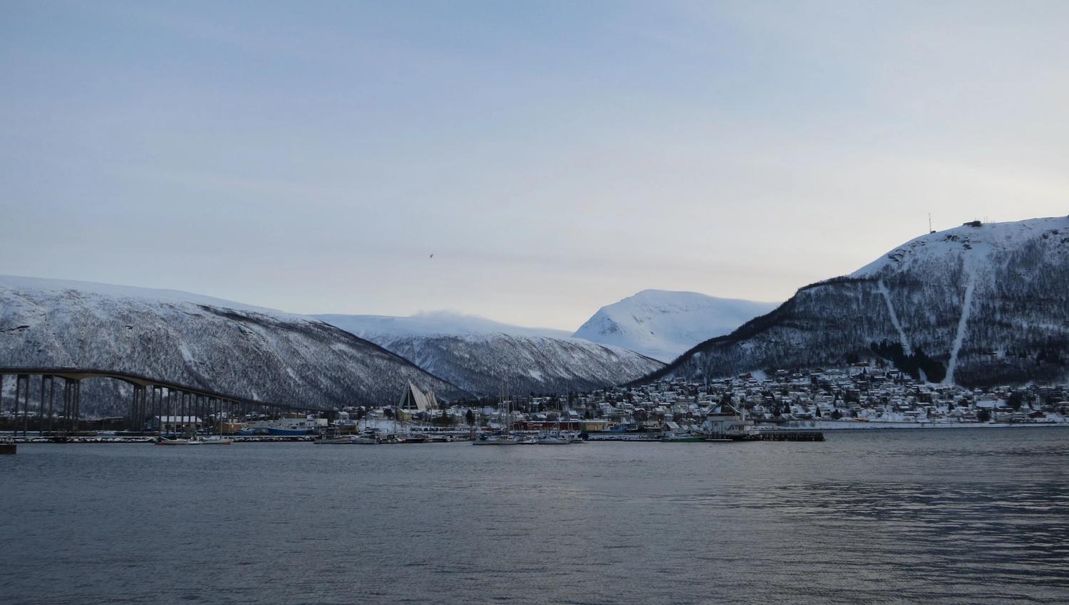 Tromsø with a bridge, buildings, and snow-covered mountains reflected in calm water.