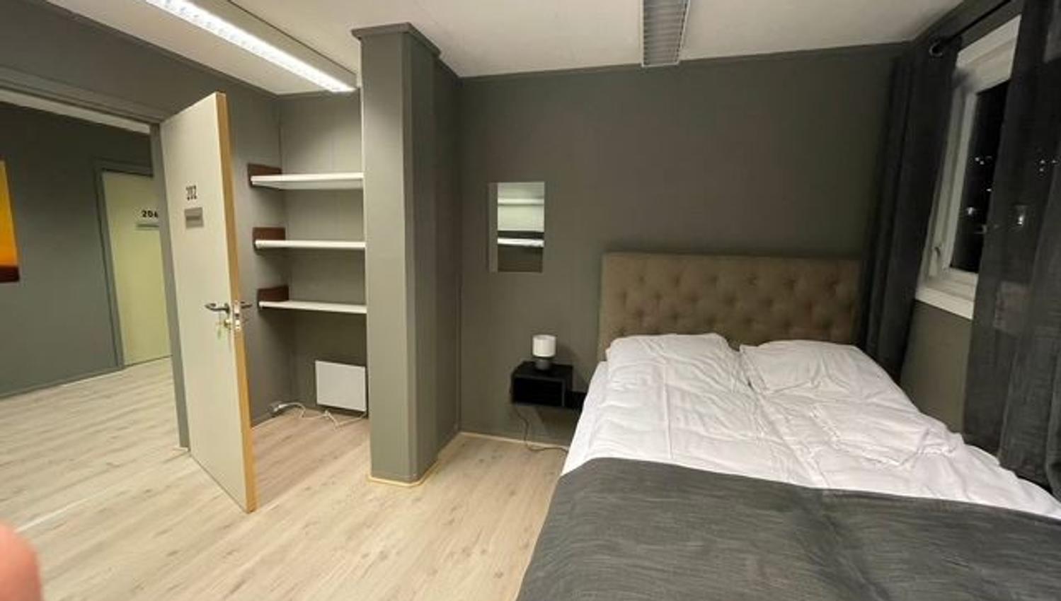 Modern bedroom with a double bed, gray walls, open shelves, bedside table, window curtains, and open door.