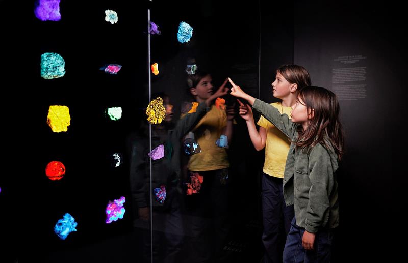 Two children point at brightly coloured minerals displayed in a dark museum room.
