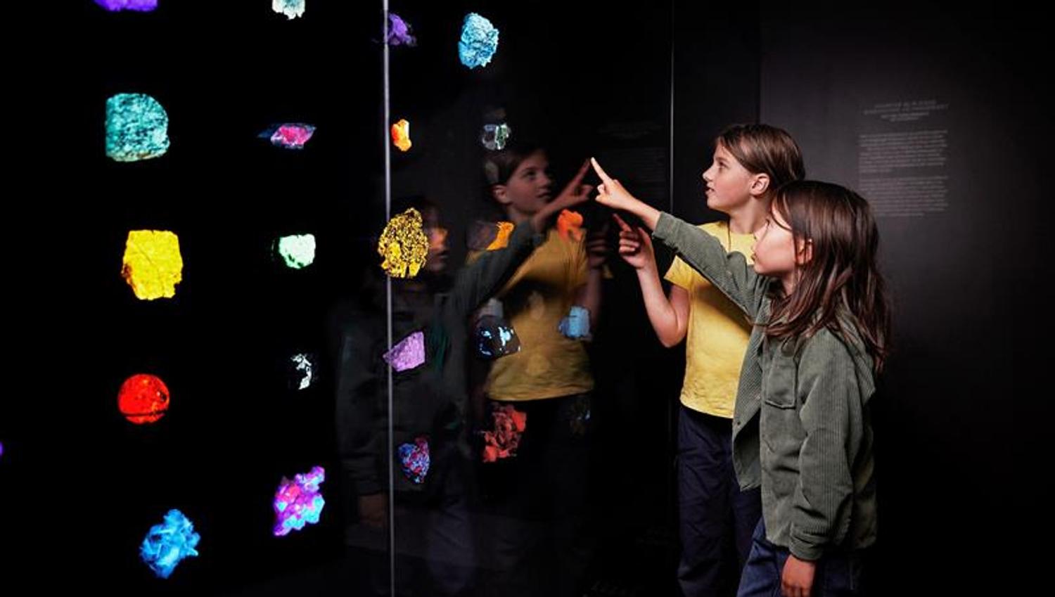 Two children point at brightly coloured minerals displayed in a dark museum room.