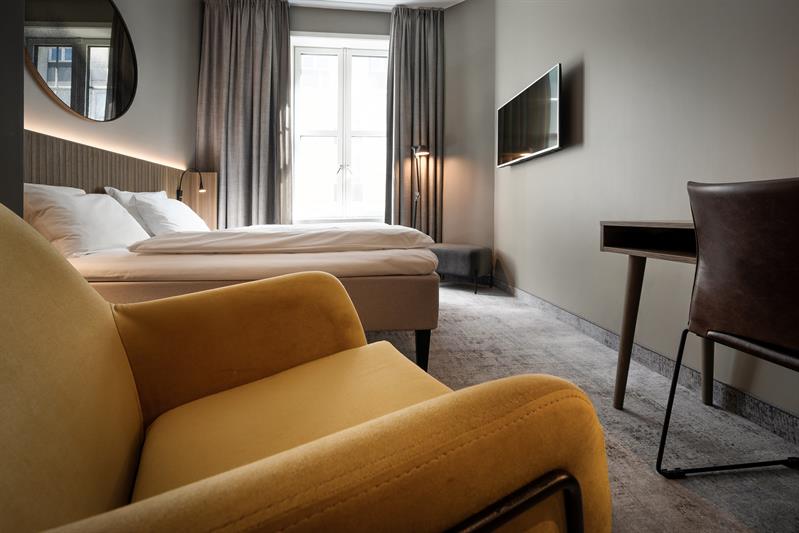 Standard room with a yellow armchair, large double bed, desk and a TV screen on the wall.