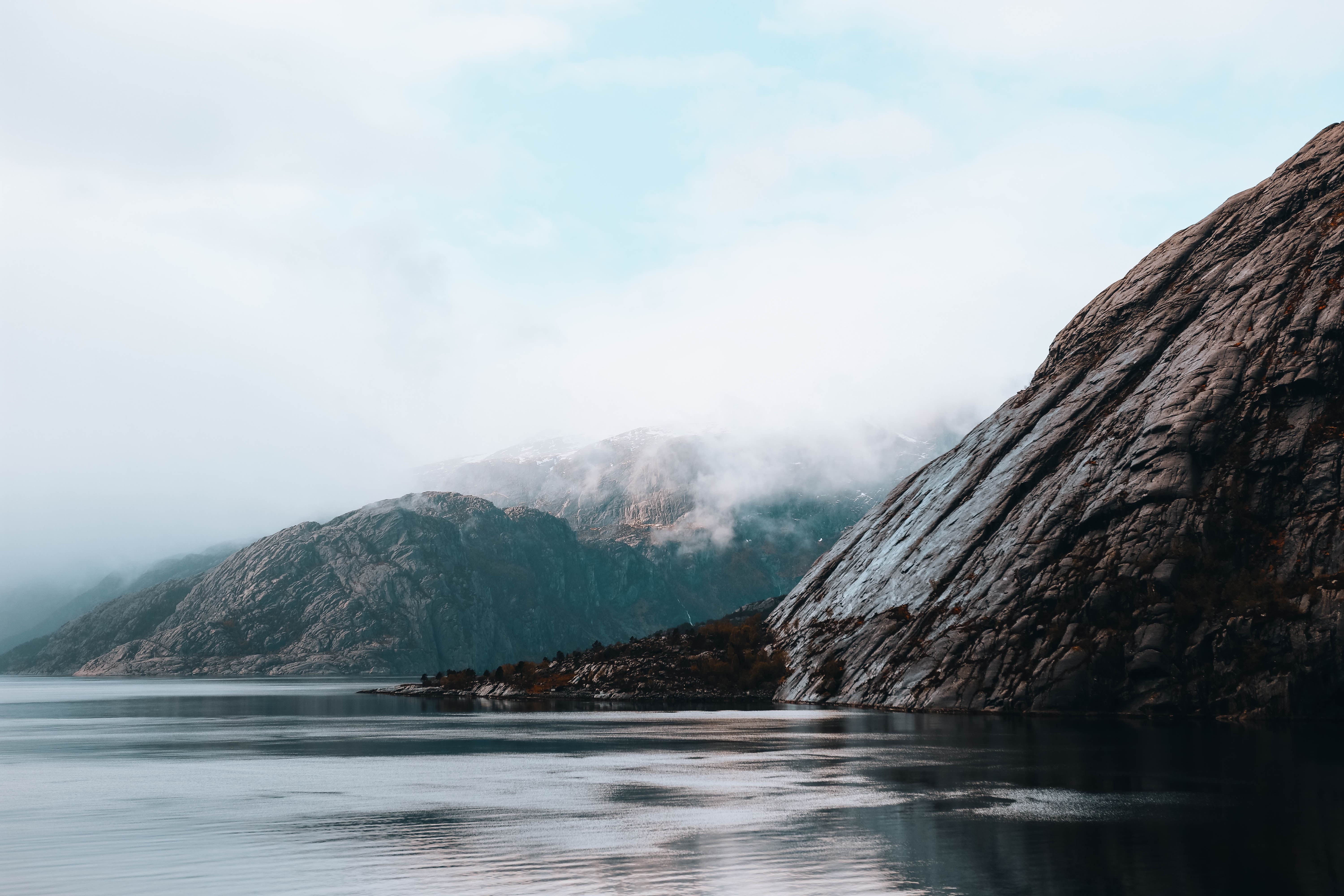 Calm water beside steep rocky mountains, with low clouds and fog drifting across the distant ridgelines.
