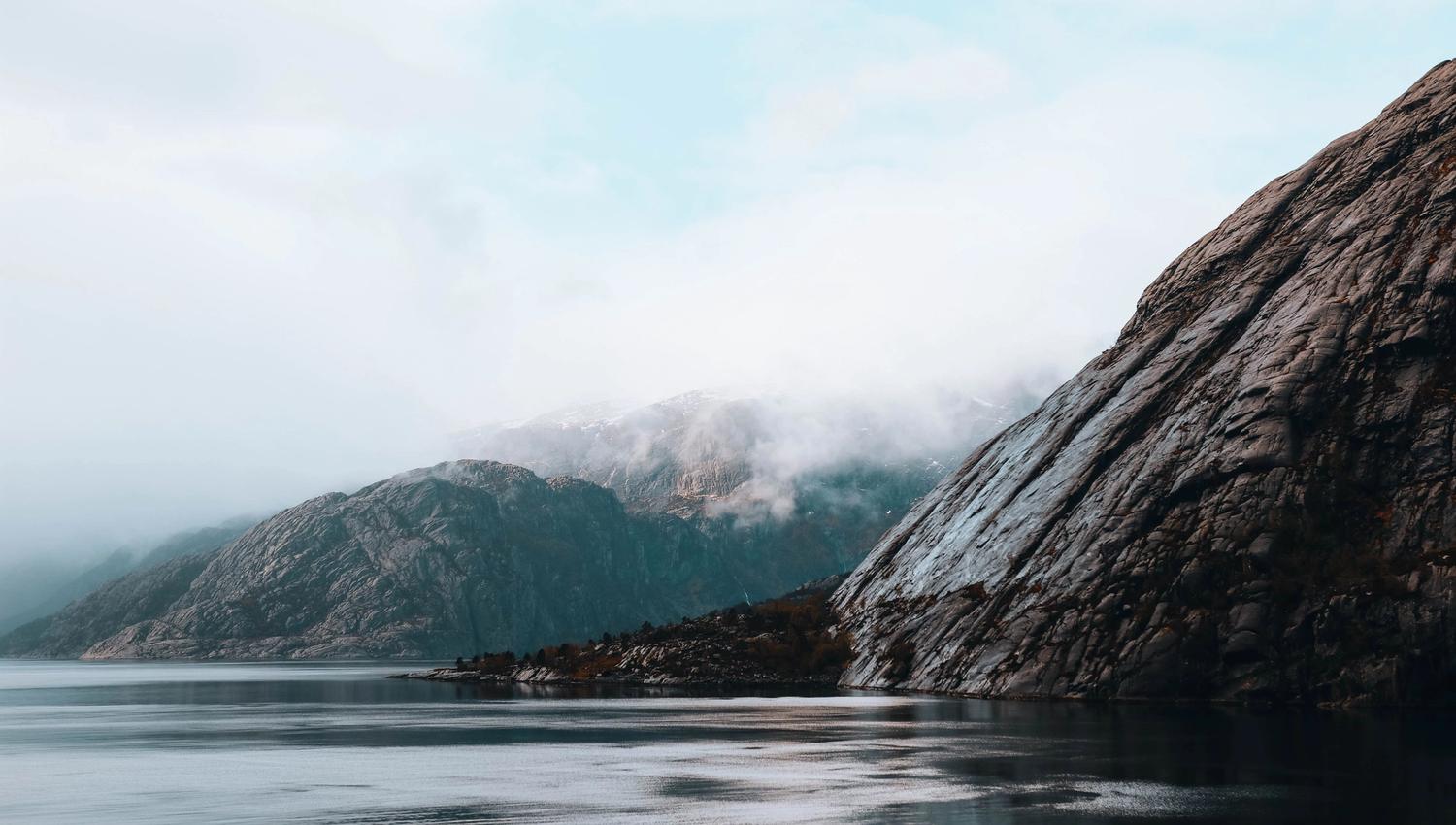 Calm water beside steep rocky mountains, with low clouds and fog drifting across the distant ridgelines.