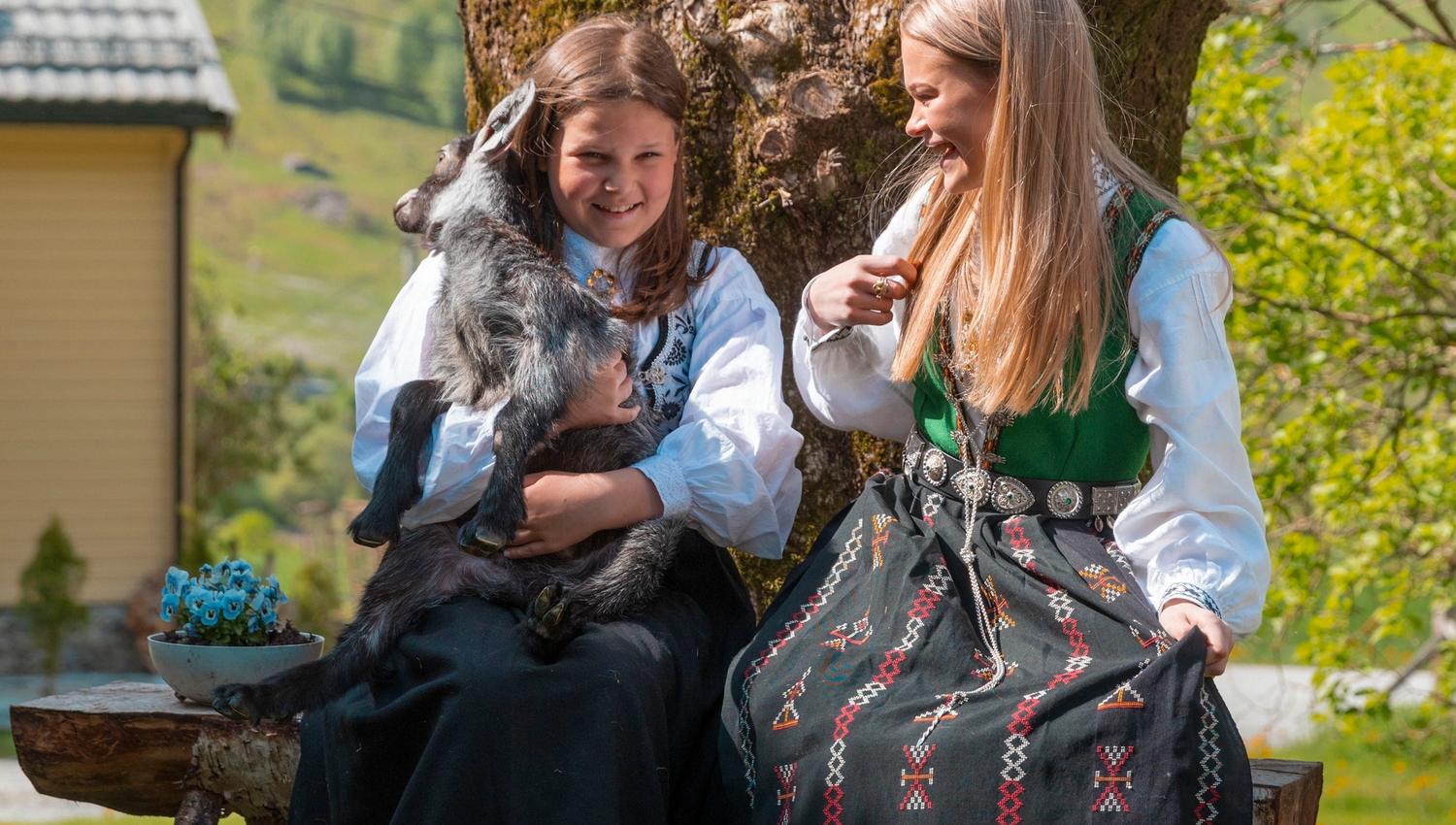 Two women in traditional dresses sit on a bench by a mossy tree, one holding a goat, in a sunny hillside setting.