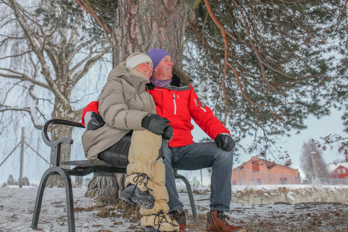To personer sitter på en benk ved et tre i et snødekt område.